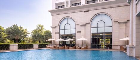 A swimming pool with lounge chairs and umbrellas, surrounded by a building with large arched windows and balconies.