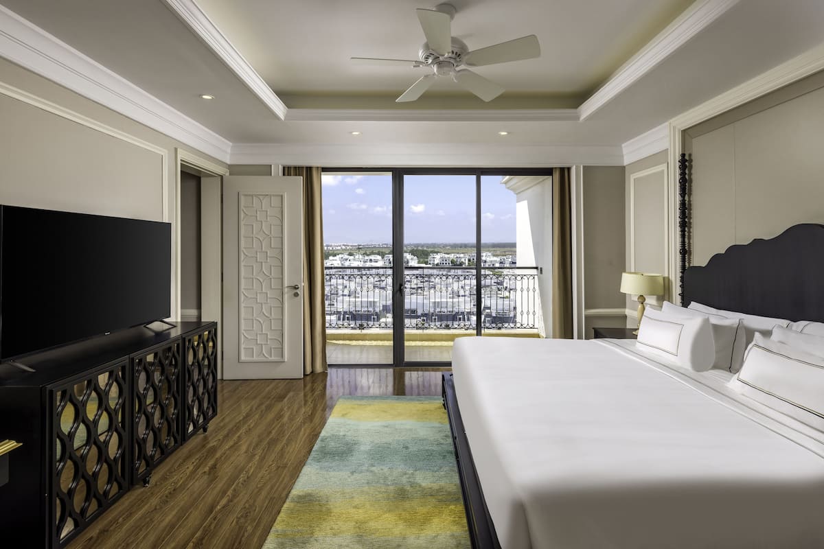 A hotel room with a large bed, a TV, a balcony with a city view, and a ceiling fan.