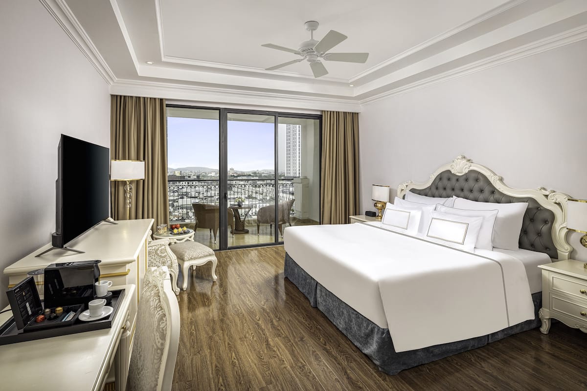 A hotel room with a large bed, a flat-screen TV, a desk with a coffee maker, and a view of the city through the balcony doors.