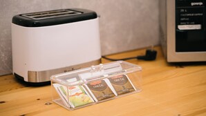 Een keuken met een broodrooster, een magnetron en een doorzichtige plastic bak met theezakjes.