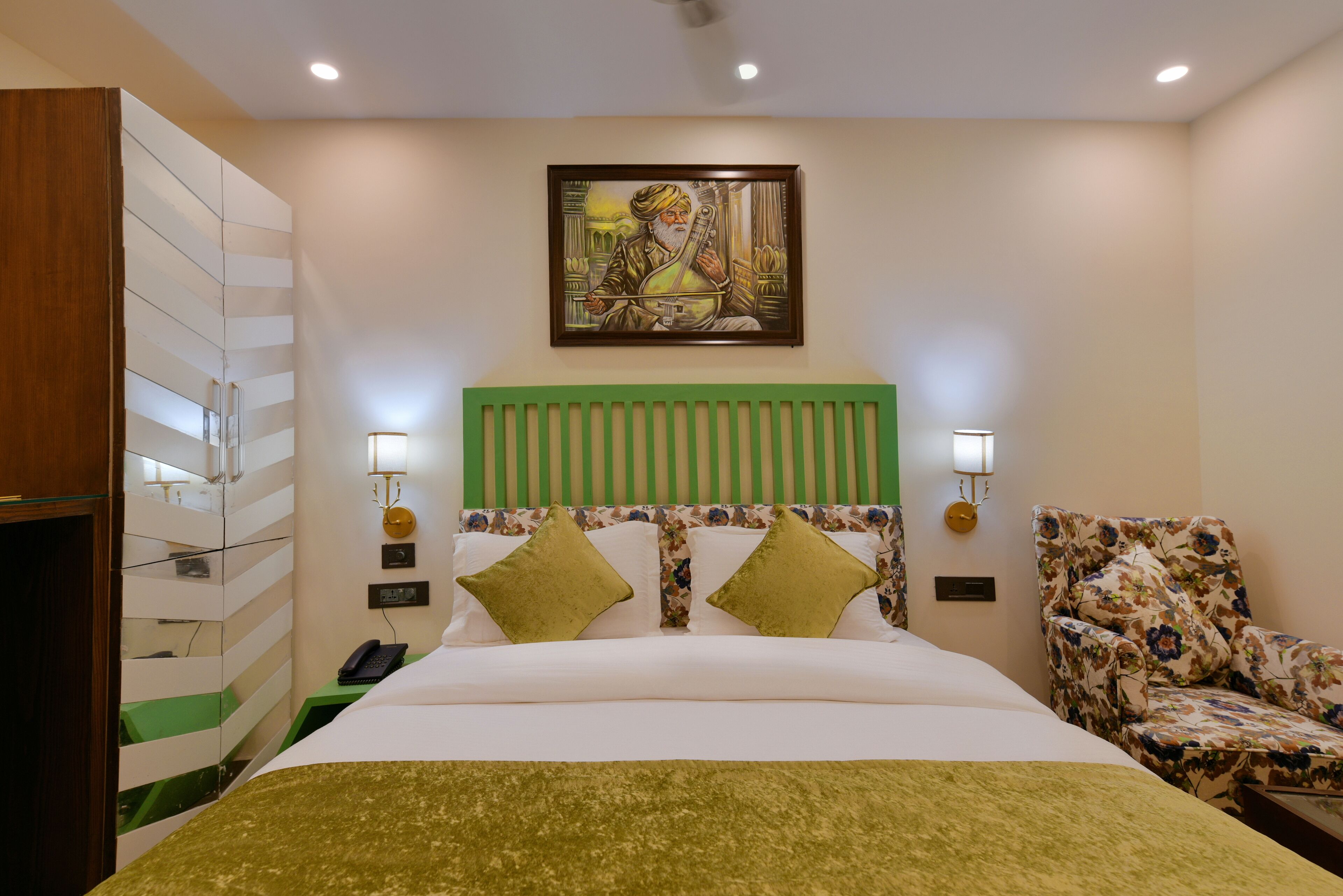 A hotel room with a bed, a green headboard, two green pillows, a floral armchair, a painting on the wall, and a green carpet.