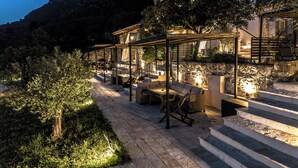 Un sentiero in pietra conduce a una zona salotto all'aperto coperta, con tavoli e sedie, immersa nel verde e con montagne sullo sfondo.