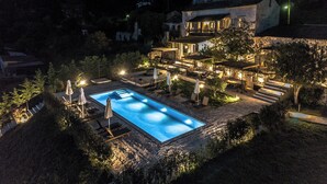 Vista notturna di una zona piscina con terrazza in pietra, poltrone relax e un edificio a più piani con illuminazione esterna.
