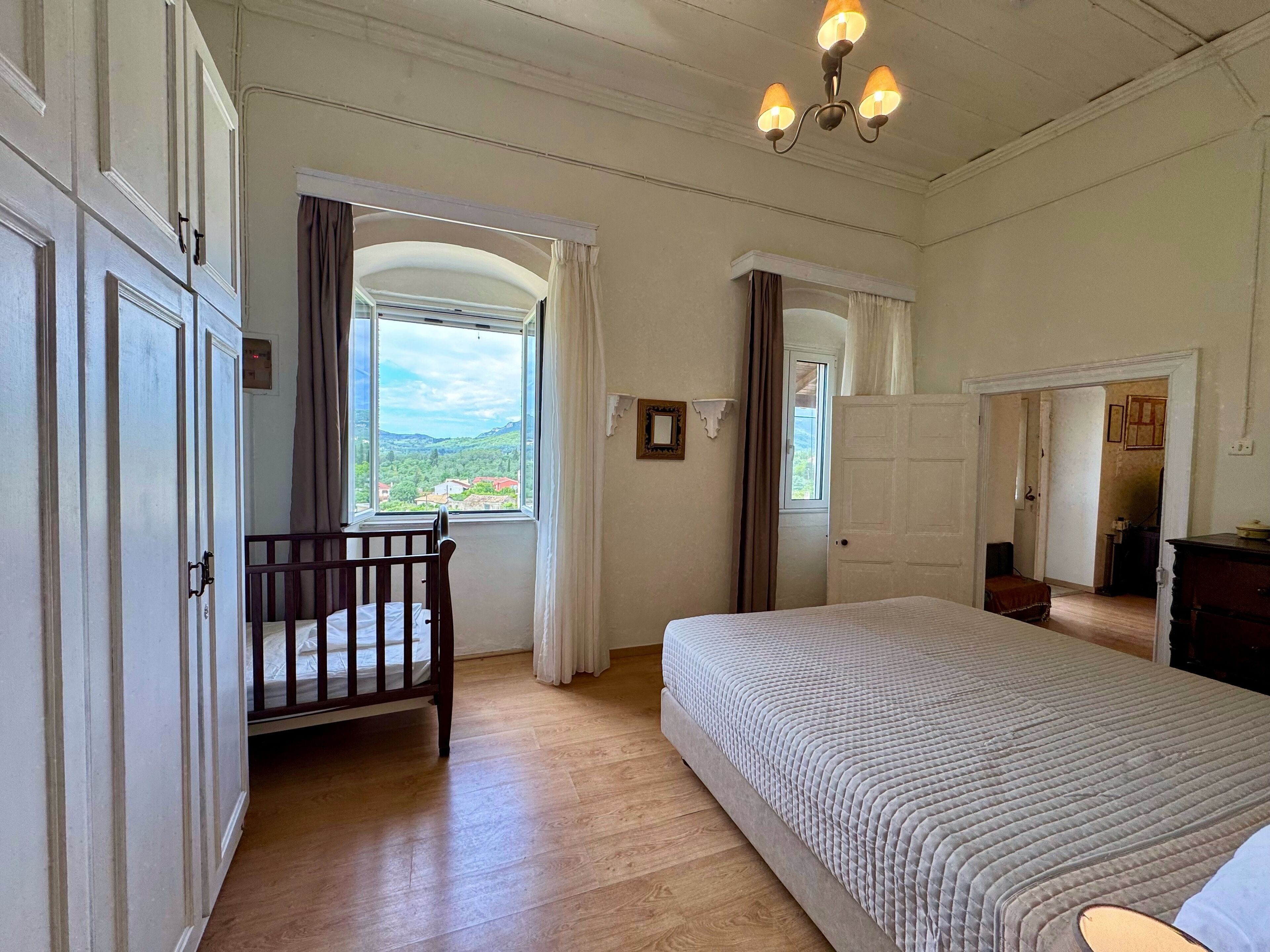 Een slaapkamer met een groot bed, een houten kinderbedje, een raam met uitzicht op het landschap, en een kroonluchter.