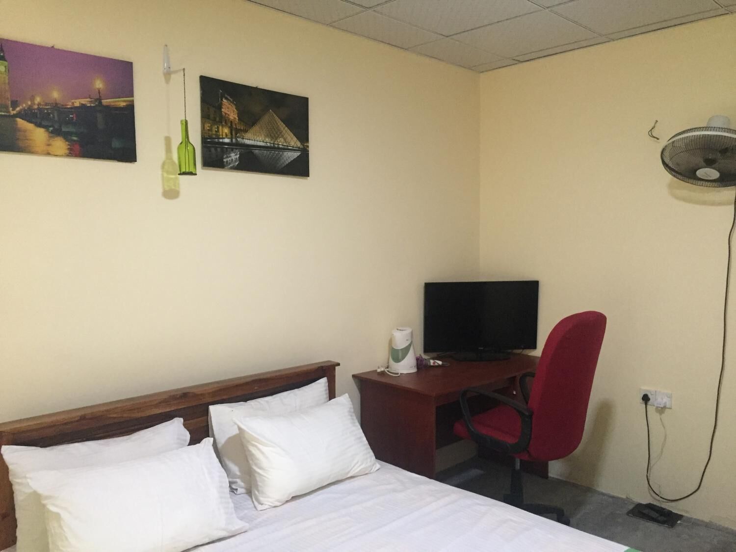 A hotel room with a bed, a desk with a television, a red chair, and two paintings on the wall.