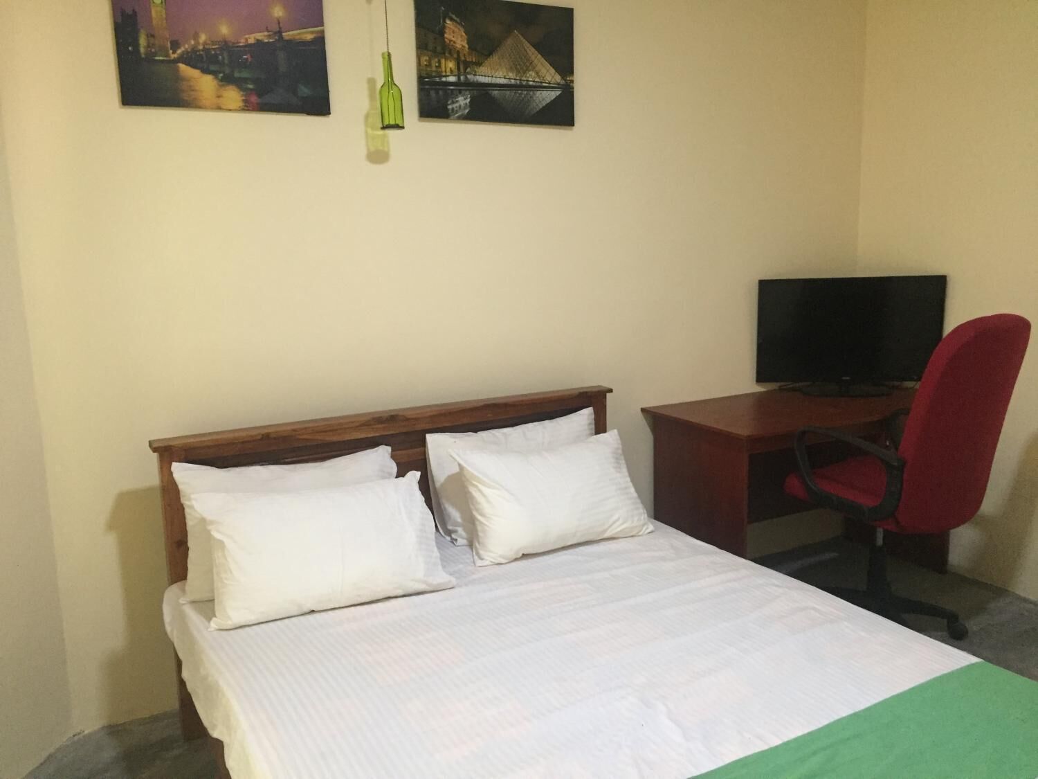 A bed with white bedding, a wooden headboard, and two pillows. A desk with a red office chair and a television is to the right.