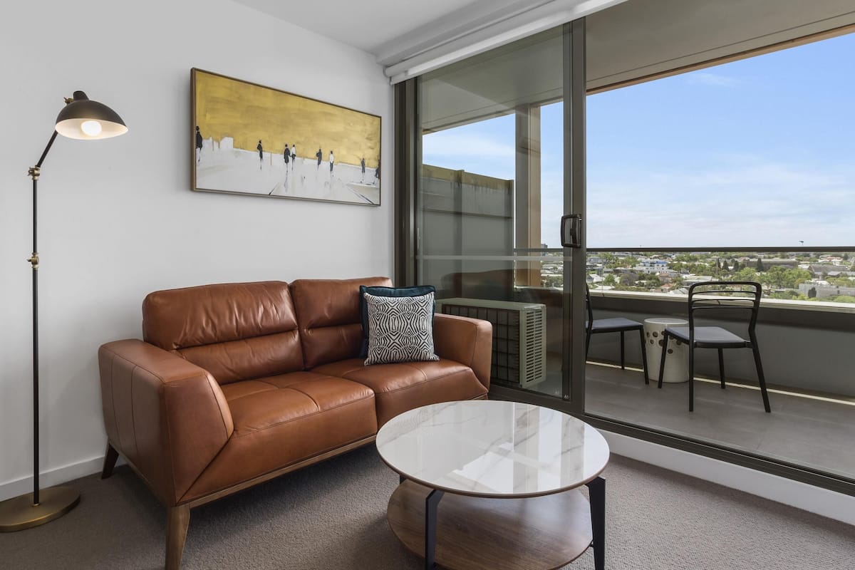 A modern living room with a leather sofa, a glass table, and a city view through large windows.