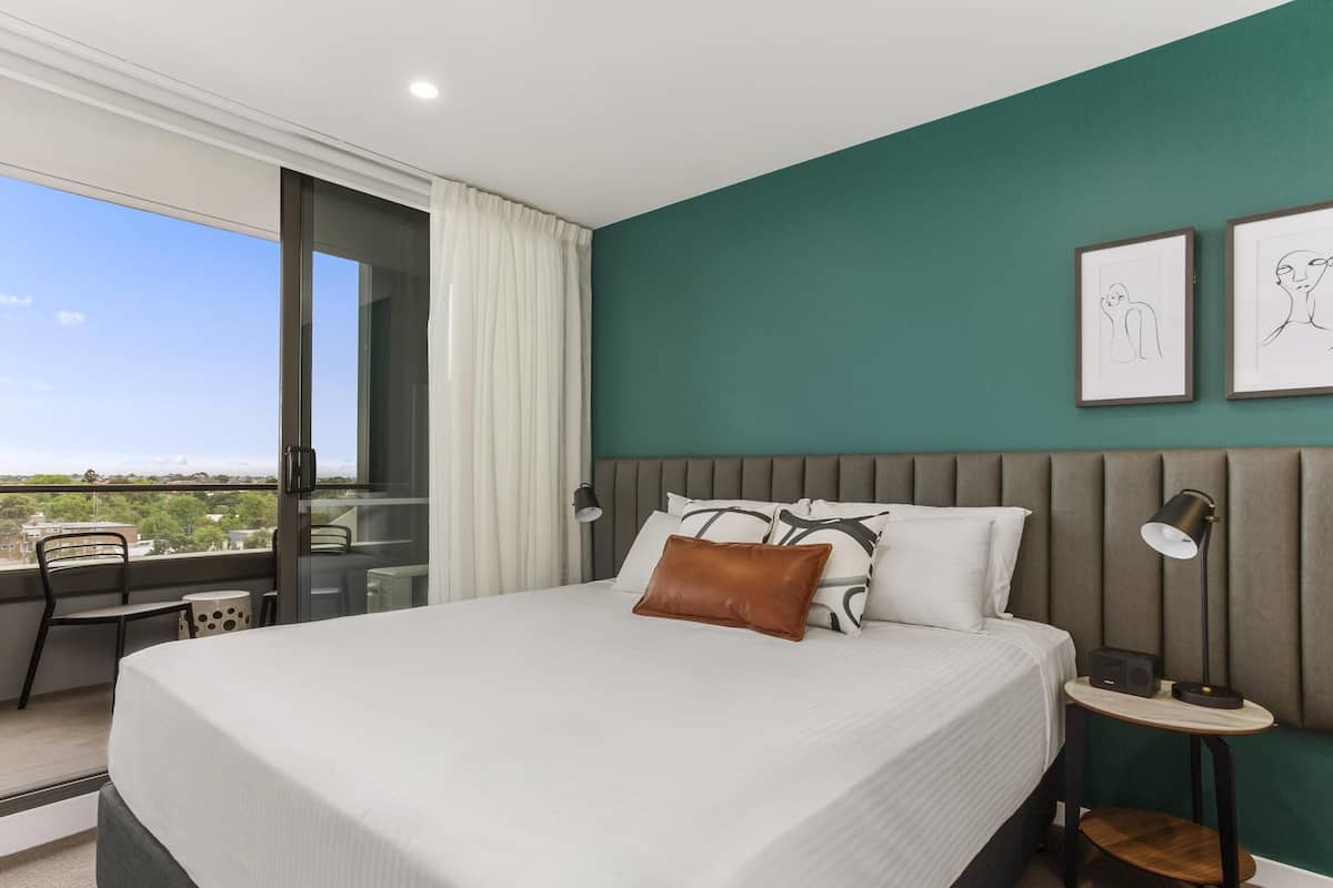 A modern hotel room with a large bed, a view through a sliding glass door, a small table with a chair, and two framed artworks on the wall.