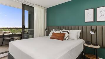 Une chambre d’hôtel moderne avec un grand lit, une vue dégagée sur l’extérieur grâce à une porte-fenêtre coulissante, une petite table avec une chaise et deux œuvres d’art encadrées accrochées au mur.