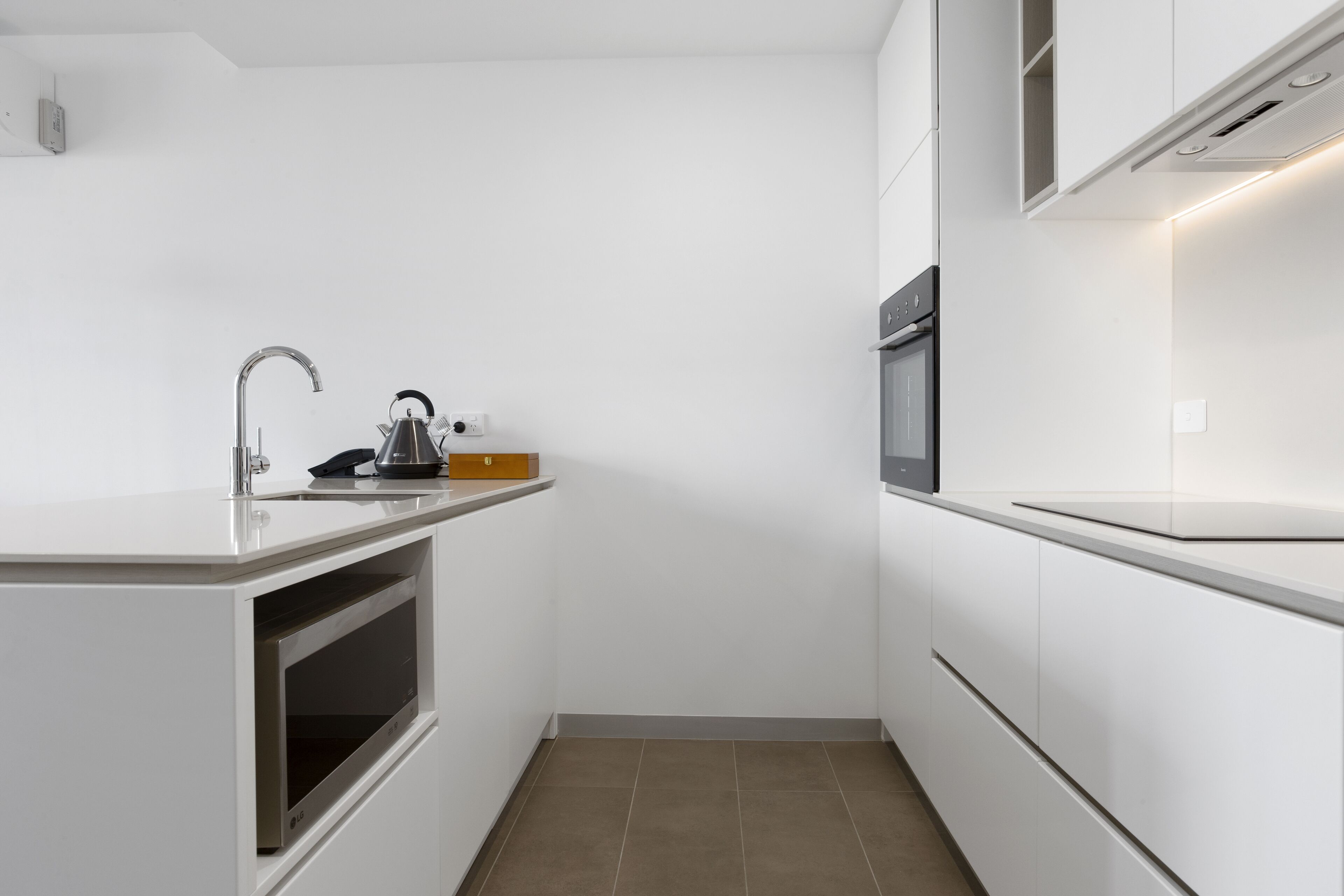 A compact kitchen with white cabinetry, a built-in oven, a stainless steel sink, and a kettle on the countertop.