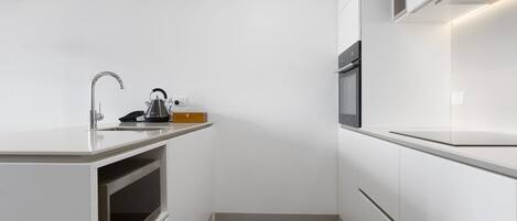 A compact kitchen with white cabinetry, a built-in oven, a stainless steel sink, and a kettle on the countertop.