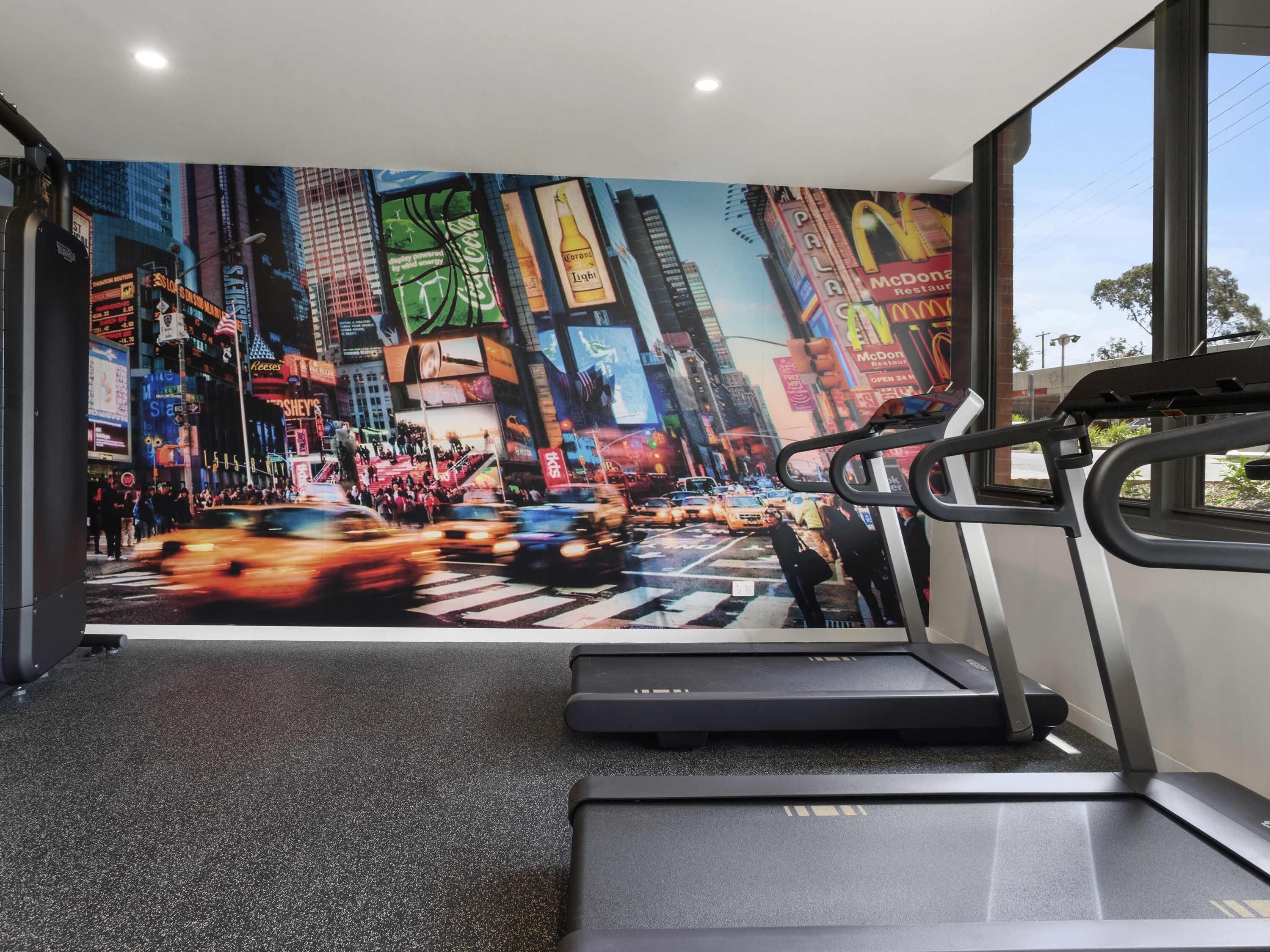 A gym with a large mural of a busy city street, a treadmill, and a window with a view of the outside.