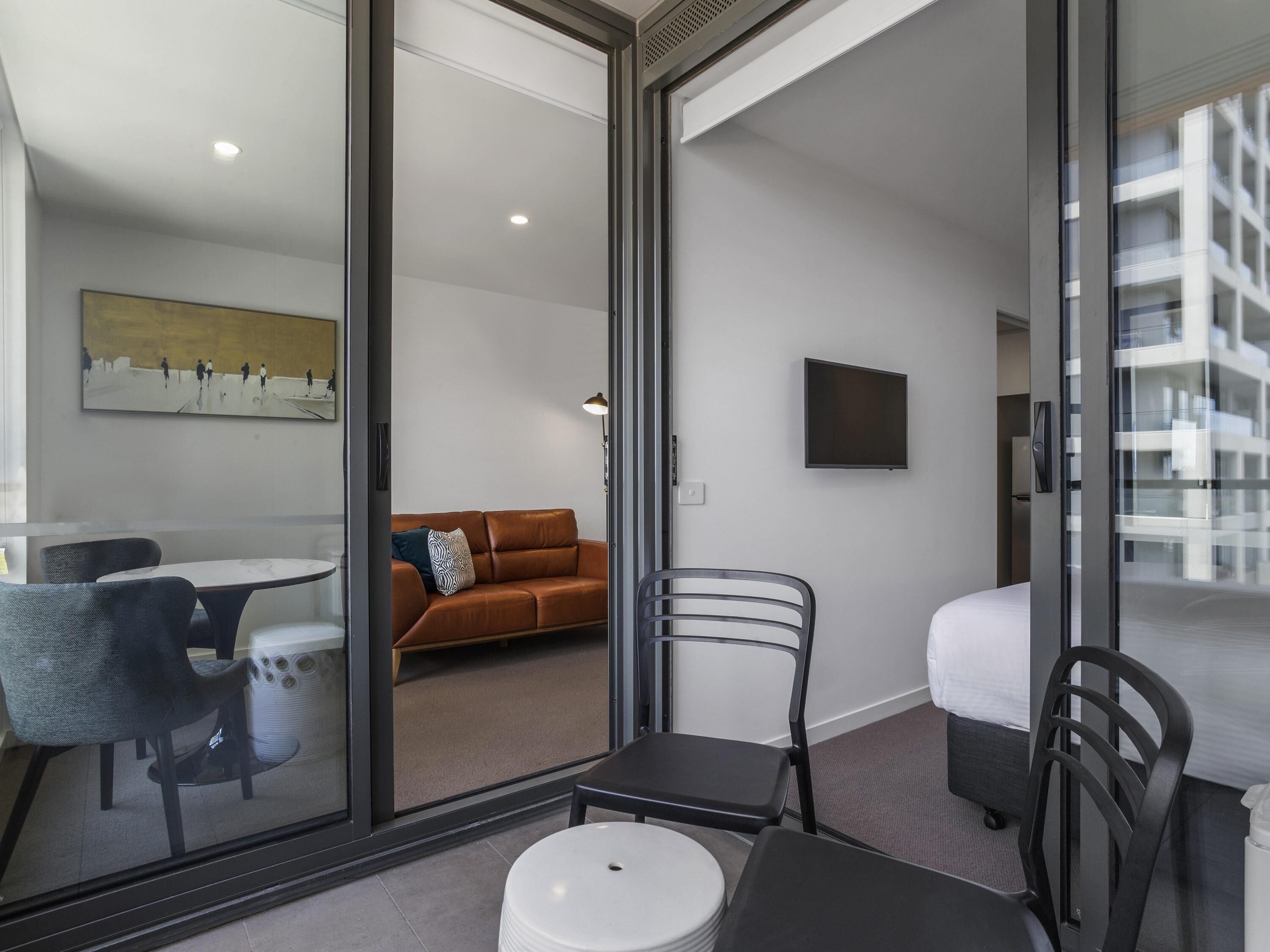 Habitació d'hotel moderna amb una porta de vidre que dona a un balcó, un sofà, una taula de dinar amb cadires i un televisor de pantalla plana fixat a la paret.