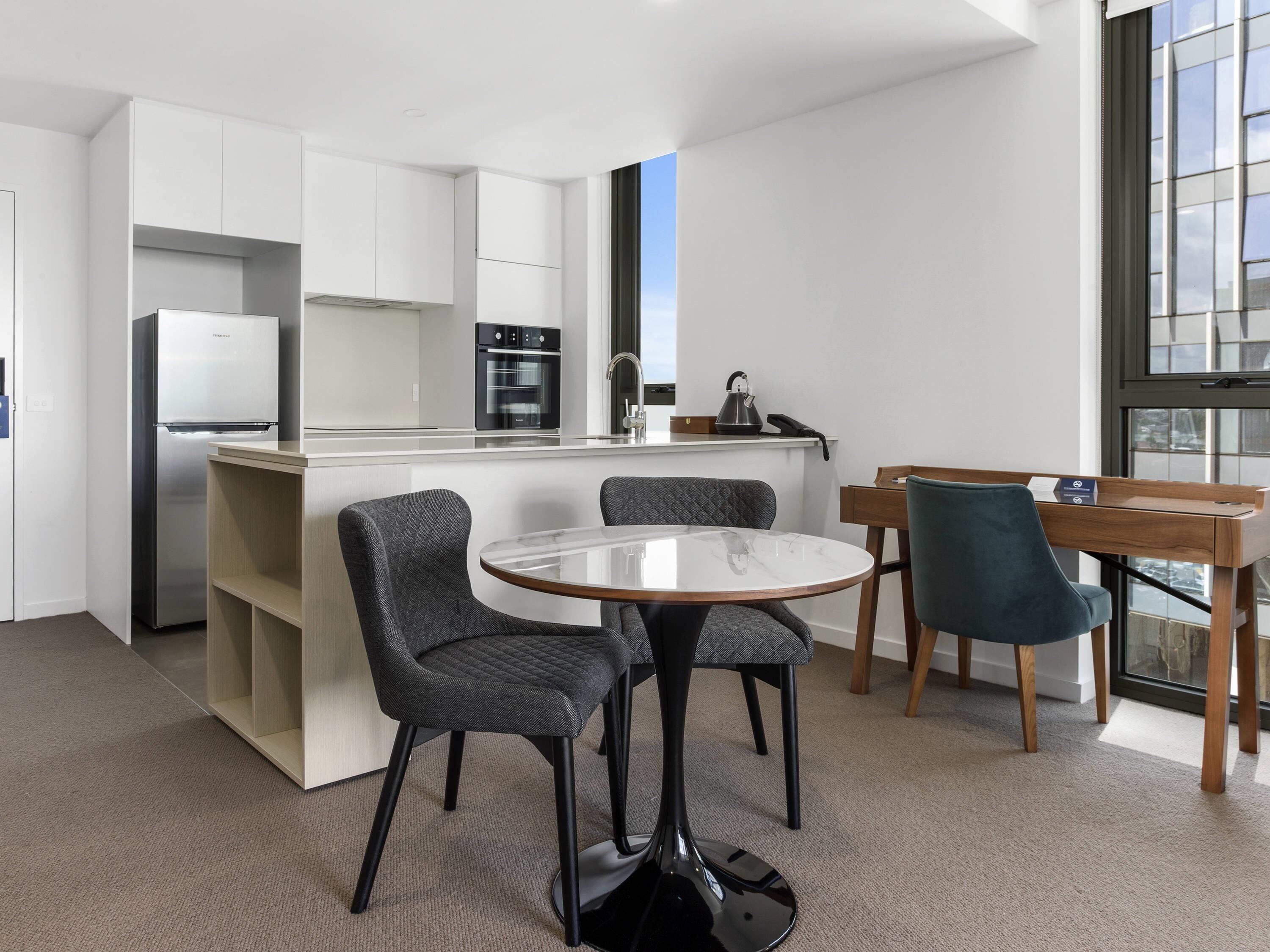 A modern kitchen with white cabinets, a stainless steel refrigerator, and a built-in oven. There is a round table with two chairs and a wooden desk with a chair.
