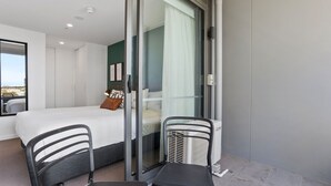 Une chambre d’hôtel moderne, équipée d’un lit, d’une porte vitrée donnant sur un balcon, d’une petite table et d’une chaise.