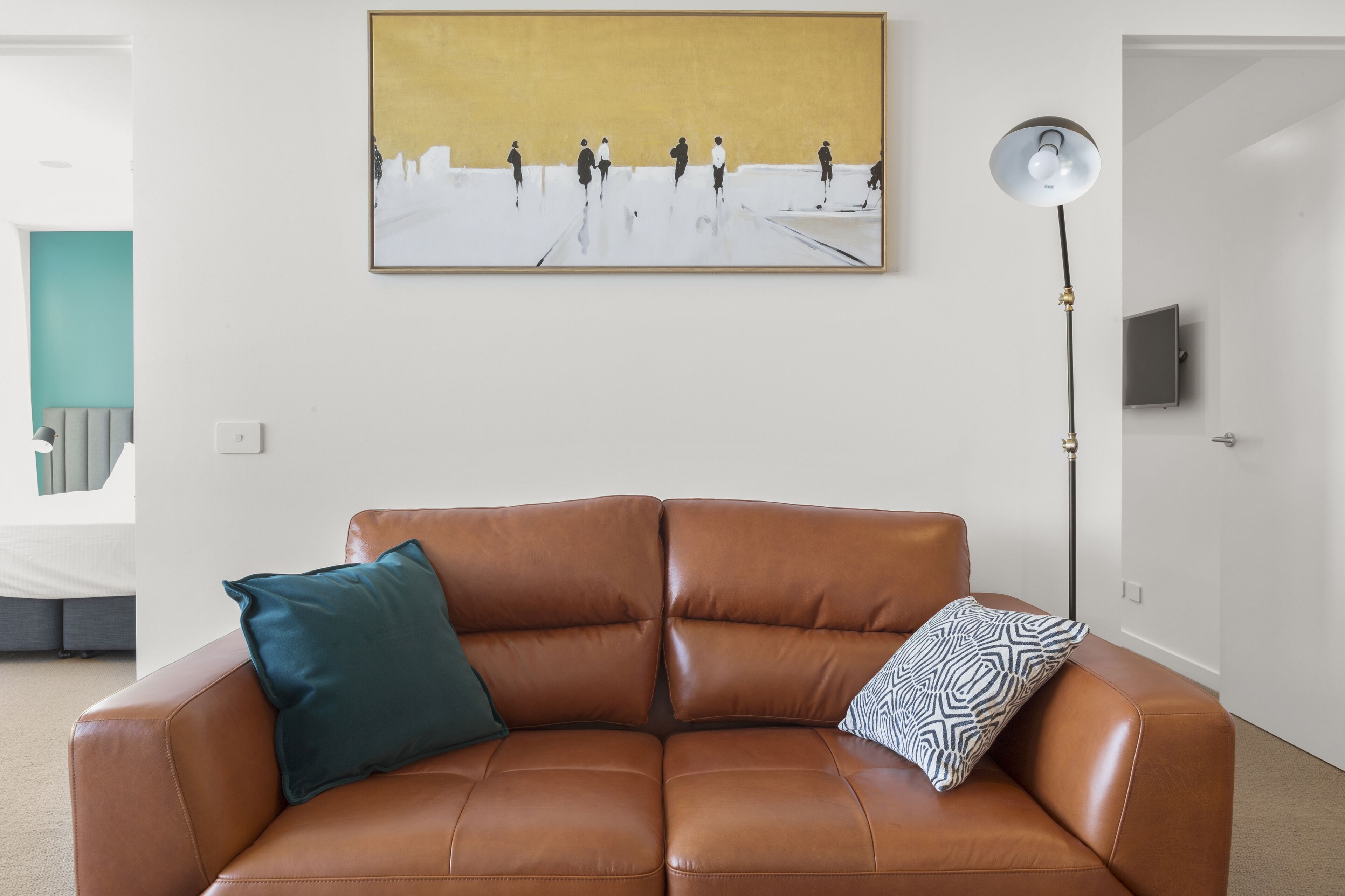 A brown leather sofa with teal and patterned pillows, a floor lamp, and a painting of people on a beach.