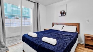 Una habitación de hotel moderna con una cama grande, un cabecero de madera, una mesita de noche y vistas a un edificio a través de un ventanal.