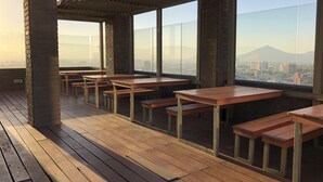 Una terraza en la azotea con mesas y bancos de madera, amplios ventanales que ofrecen vistas a la ciudad y a las montañas.