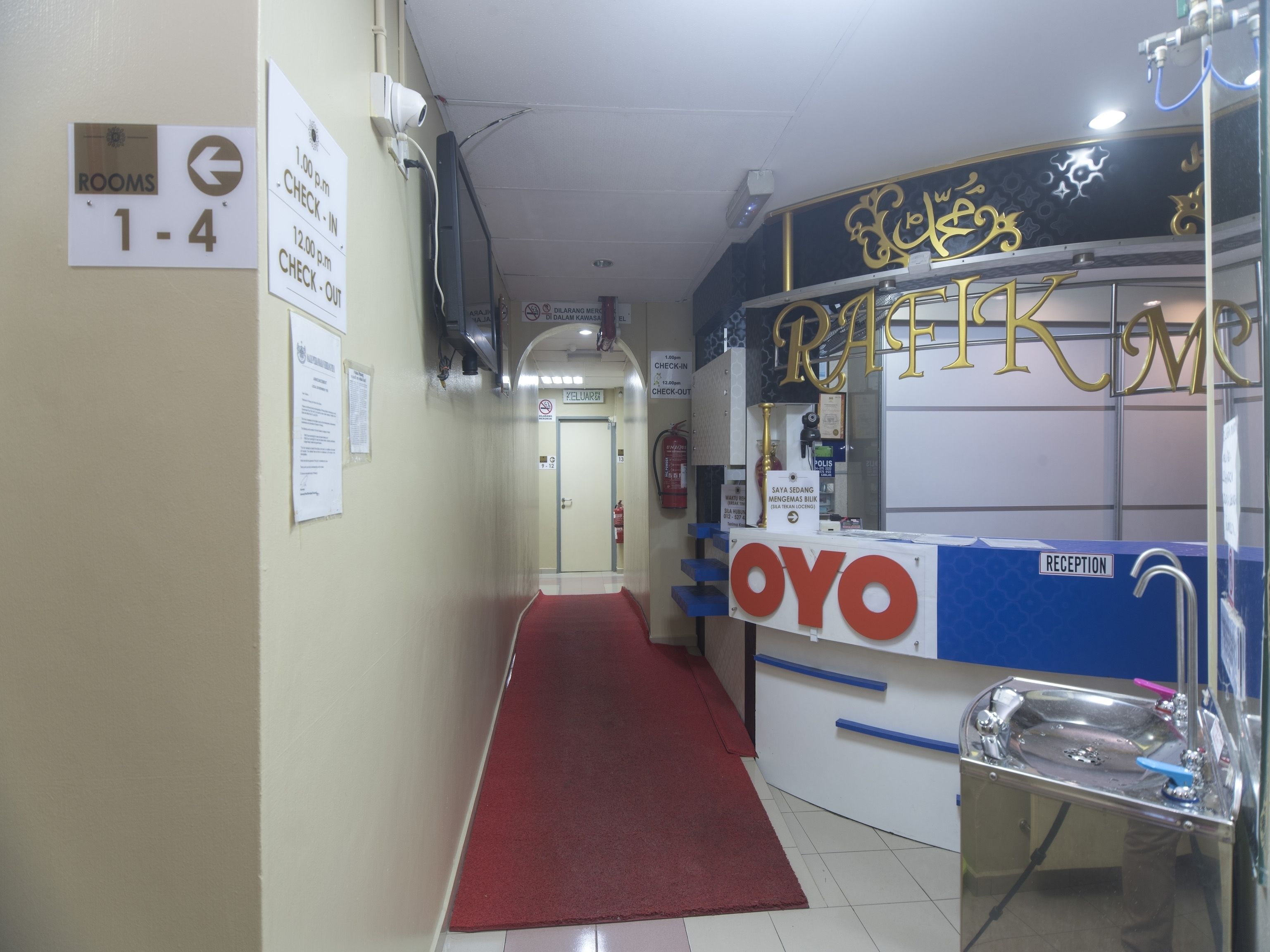 A hotel reception area with a reception desk, a red carpet, and signs indicating room numbers and check-in/out times.