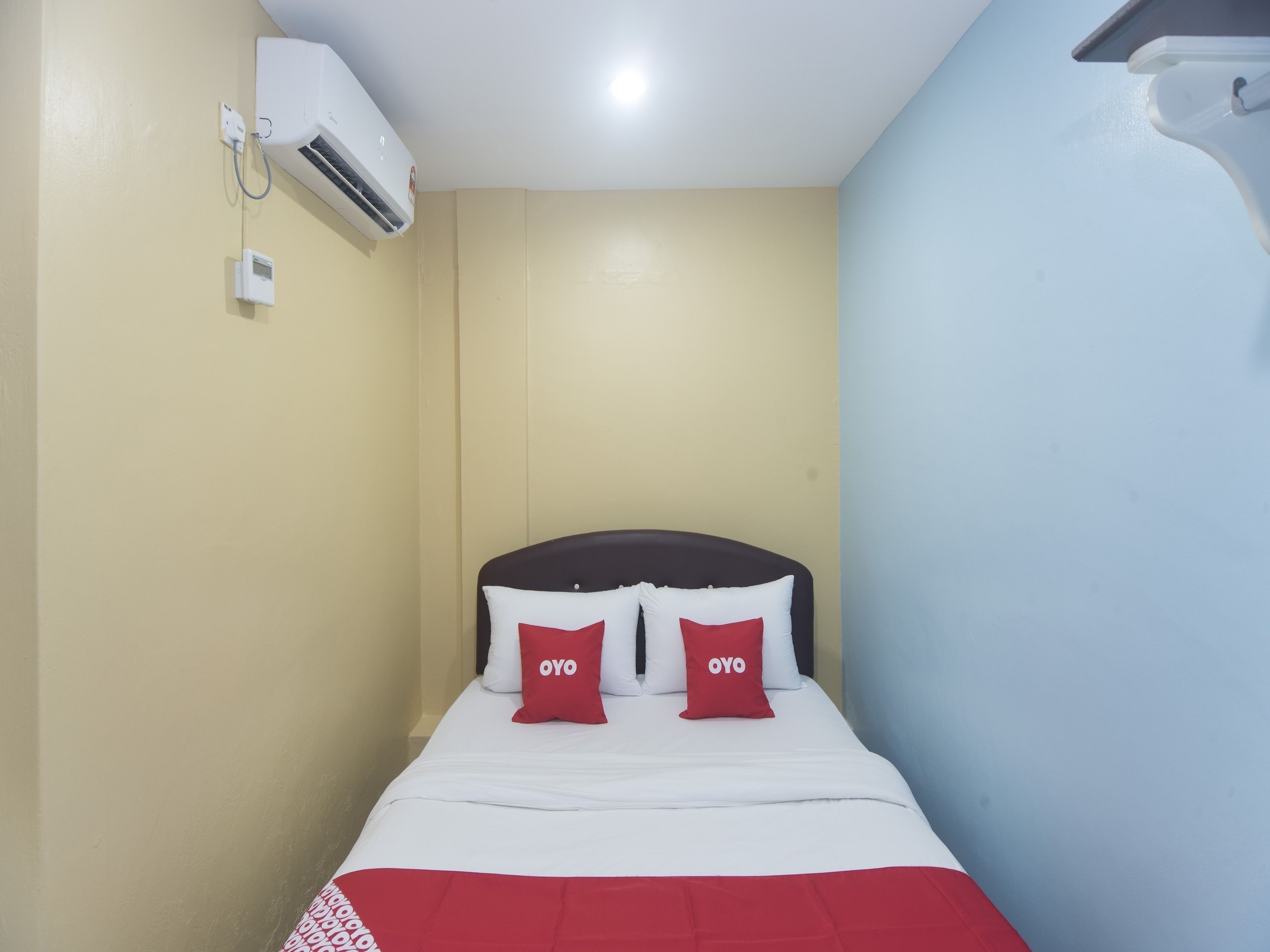 A hotel room with a bed, two pillows with the word 'OYO' on them, an air conditioning unit, and a light switch on the wall.