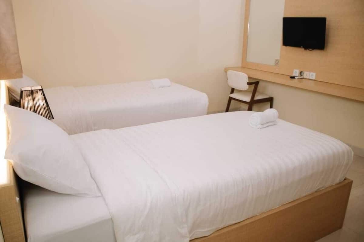 Habitación de hotel con dos camas individuales, una silla, un televisor de pantalla plana montado en la pared y una mesita de noche con lámpara.
