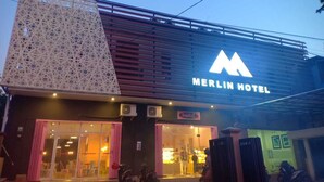 Das äußere Erscheinungsbild des Merlin Hotels mit einem gut sichtbaren 'M'-Logo, einem großen Glas Eingang und einer modernen Fassade.