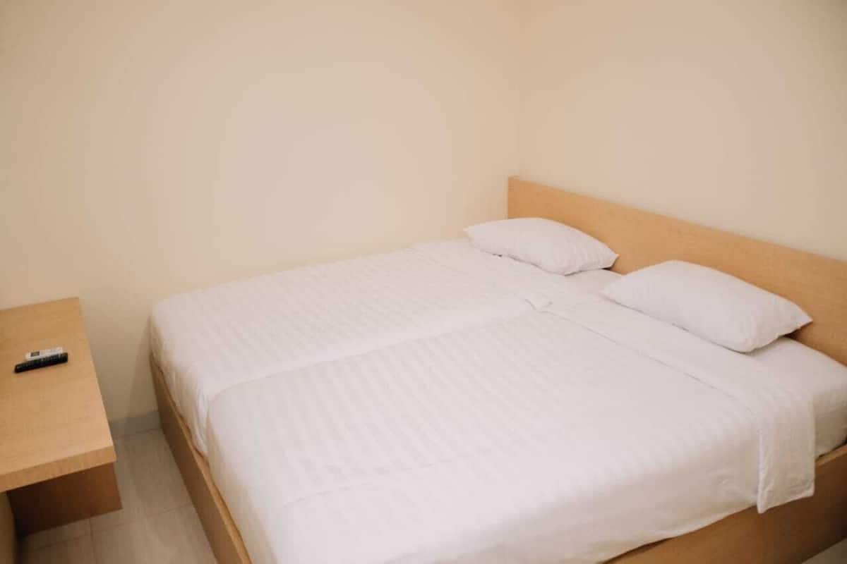 Una habitación sencilla y limpia con una cama individual, una mesita de noche de madera y un control remoto sobre la mesita.