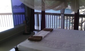 A bed with a white mattress and brown pillows, a standing fan, and a view of a rocky shoreline through a balcony with a white railing. - Hinwong Apartments Dive & Snorkel Resort (Koh Tao)