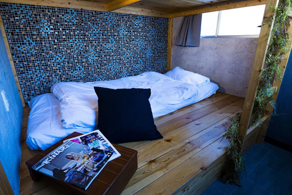 A small, cozy room with a wooden floor, a bed with white linens, a black pillow, and a magazine on a small table.