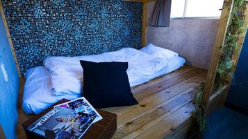 Quarto pequeno e aconchegante, com piso de madeira, uma cama com lençóis brancos, um travesseiro preto e uma revista sobre uma mesa pequena.