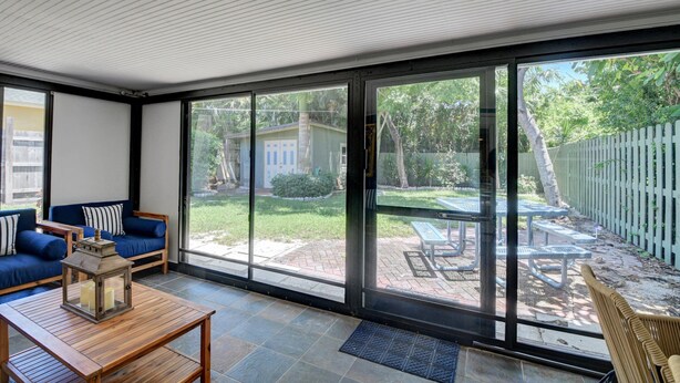 A sunlit room with a large sliding glass door leading to a patio area. Inside, there is a wooden table with a lantern, a blue sofa with striped cushions, and a wicker chair. - Gorgeous Downtown Delray Beach Cottage (Delray Beach)