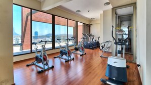 Ein geräumiges Fitnessstudio mit mehreren Trainingsgeräten, großen Fenstern und Blick auf eine Stadtlandschaft sowie einem Holzboden.