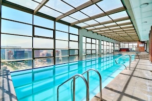 An indoor swimming pool with a city view, large windows, and a glass ceiling. - Vespucio Art (Santiago)