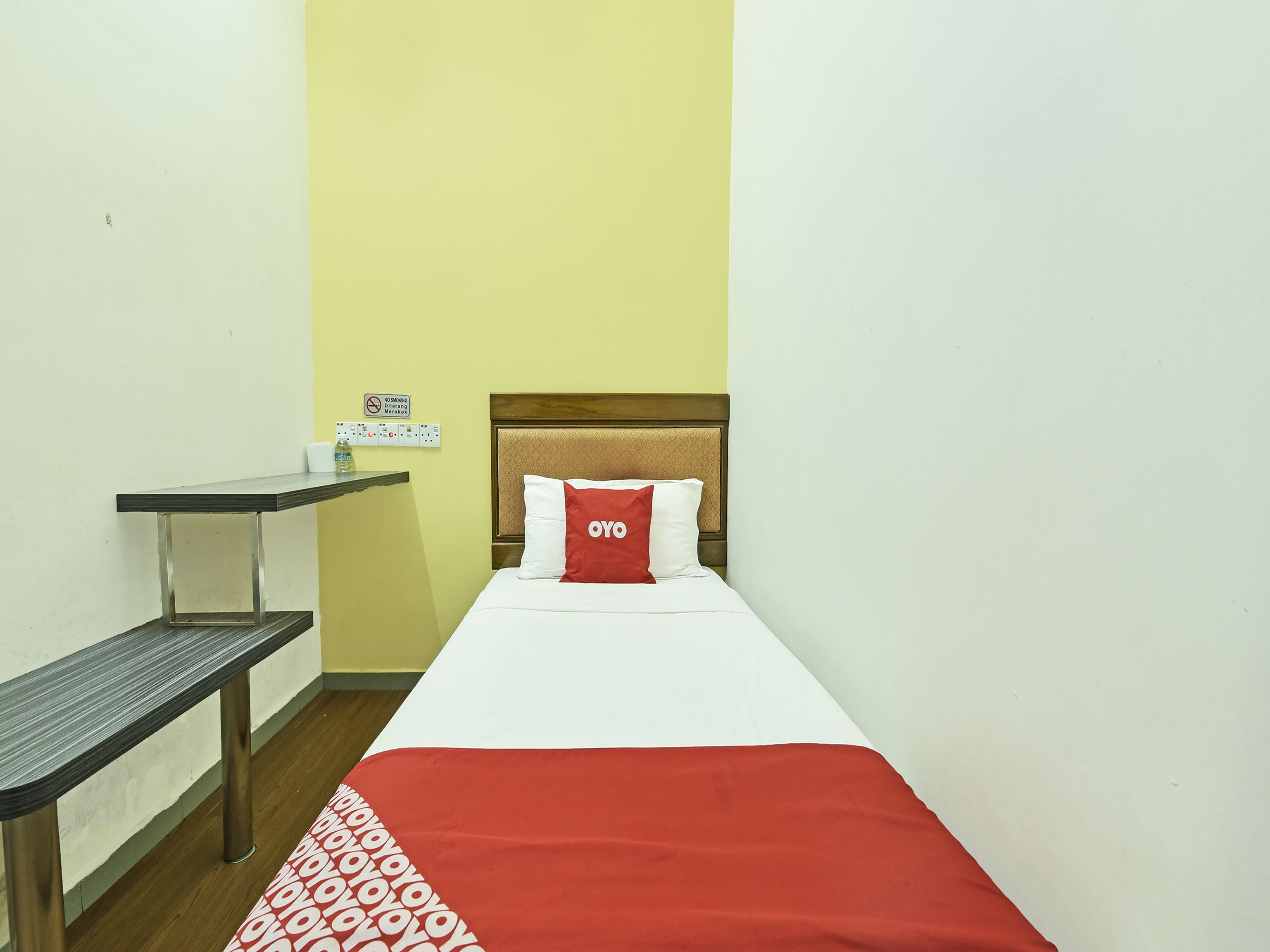 A small, single-bed room with a wooden headboard, a red and white bedspread, a small table with a chair, and a wall-mounted power outlet.