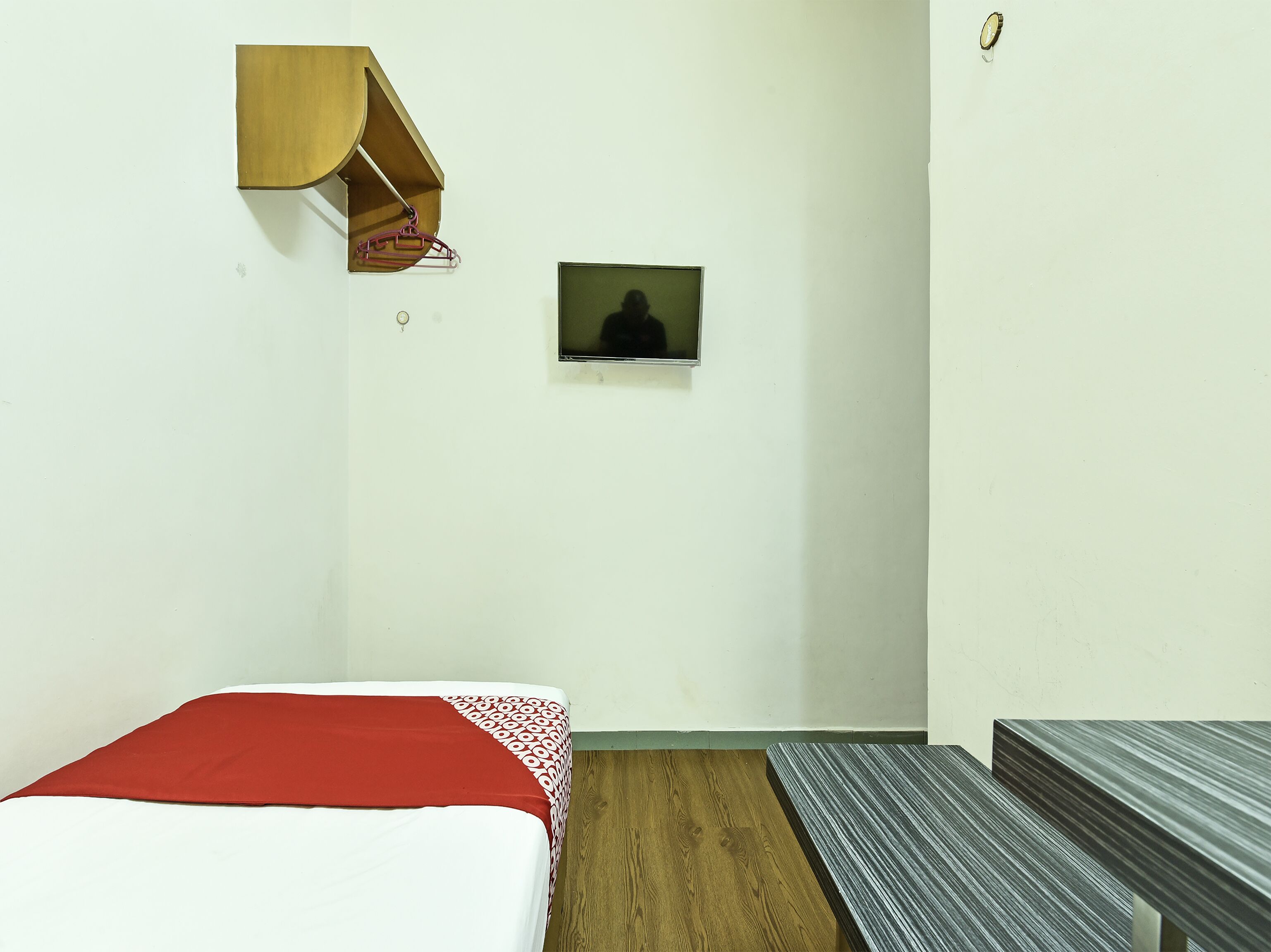 A small, sparsely furnished room with a bed, a wooden bench, a small shelf, and a television.