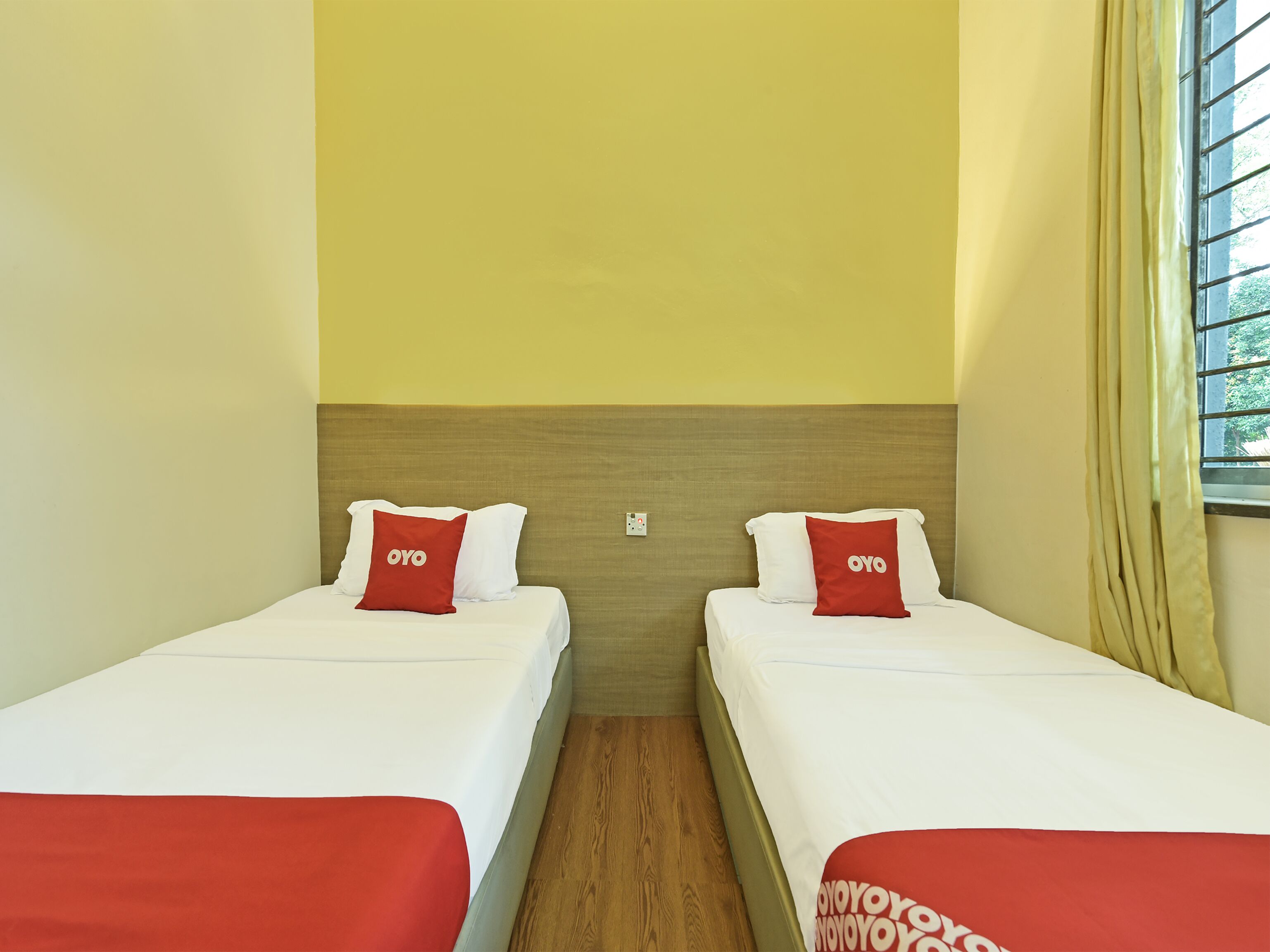 A hotel room with two single beds, each with a red pillow and a white bedspread. The room has a wooden headboard, a small window with curtains, and a wooden floor.
