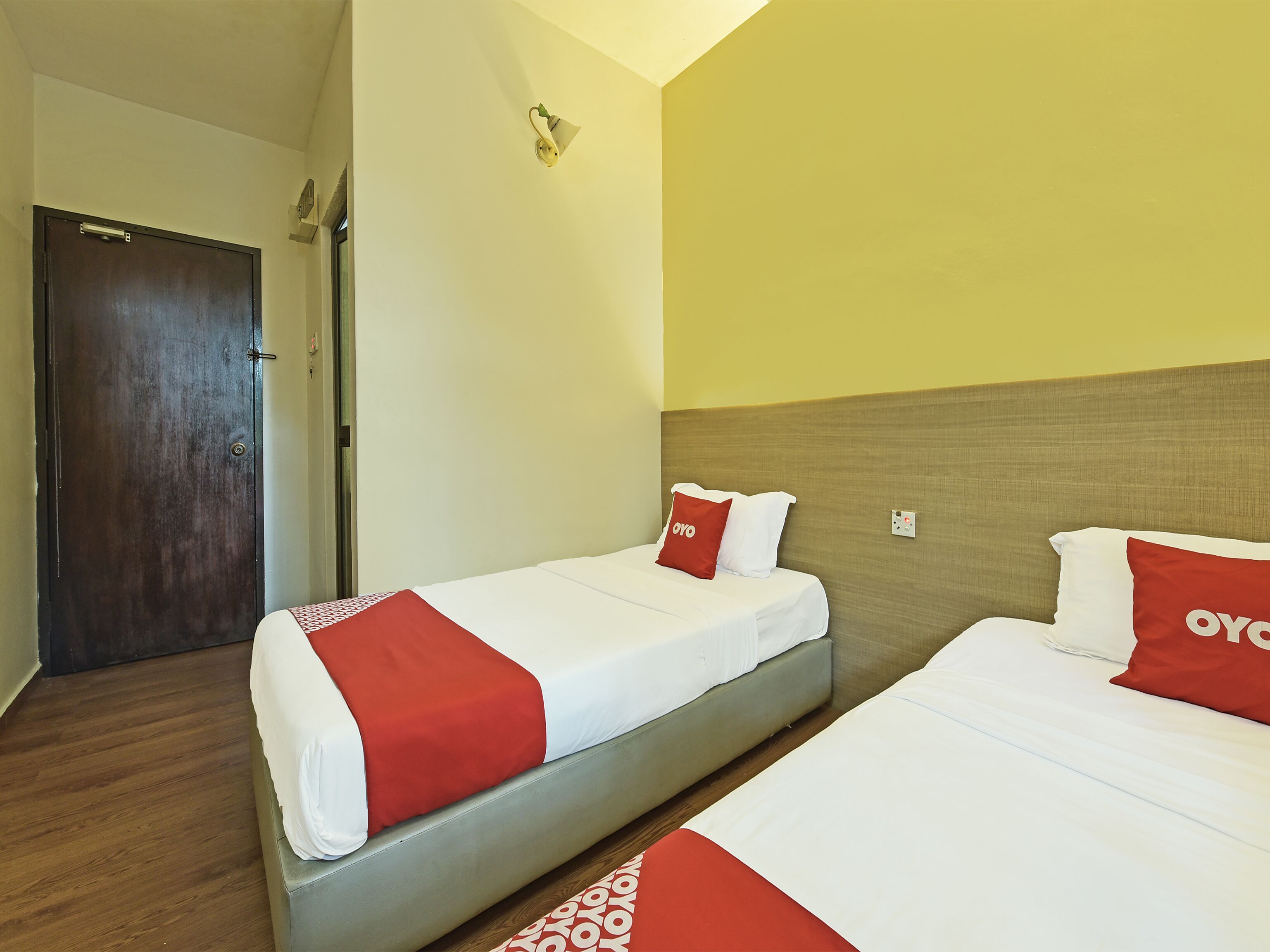 Una habitación de hotel con dos camas, cada una con ropa blanca y detalles en rojo, una puerta de madera y un interruptor de luz en la pared.