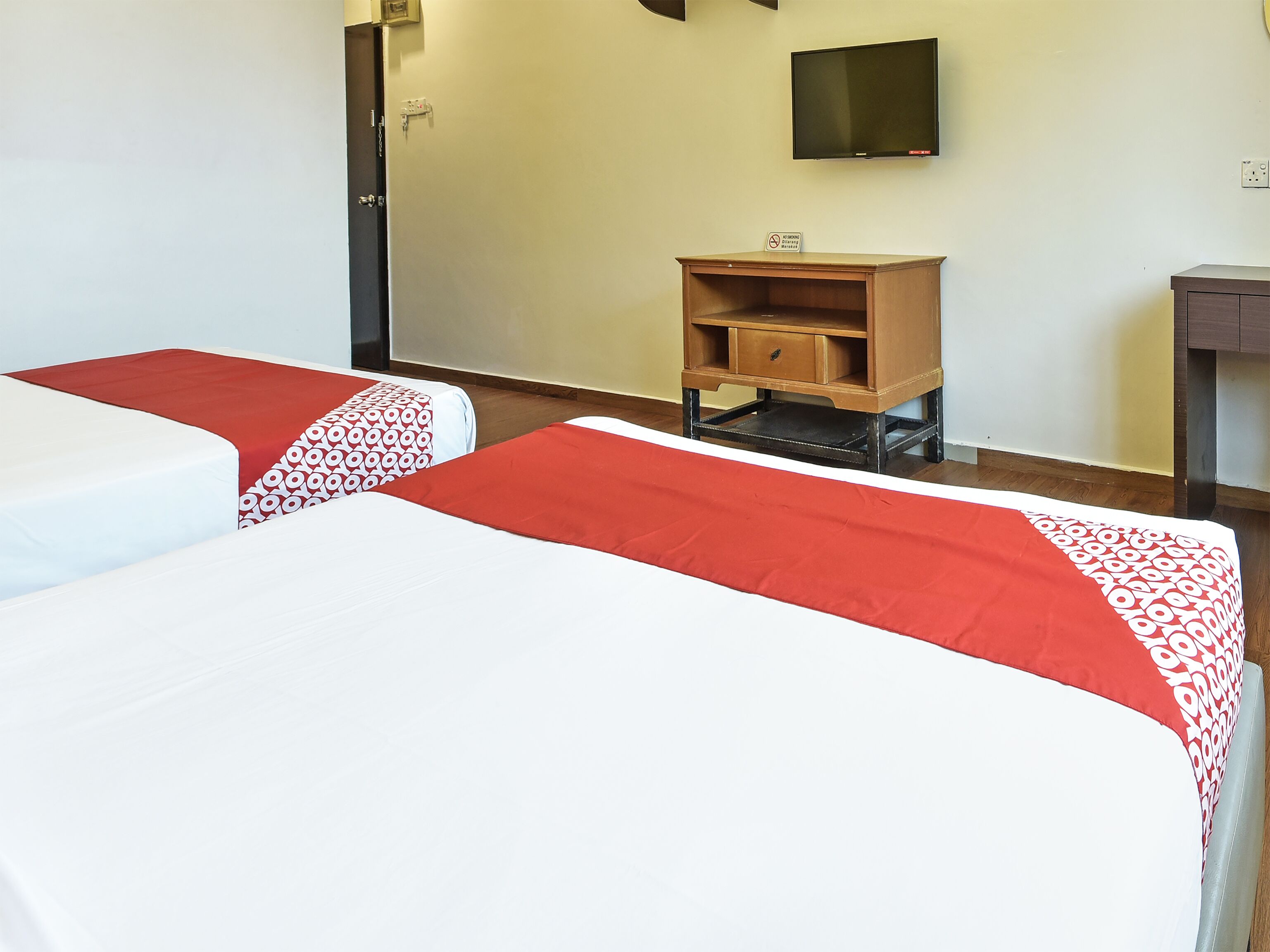 A hotel room with two beds, each with a red and white bedspread, a small wooden nightstand, a television mounted on the wall, and a door with a lock.