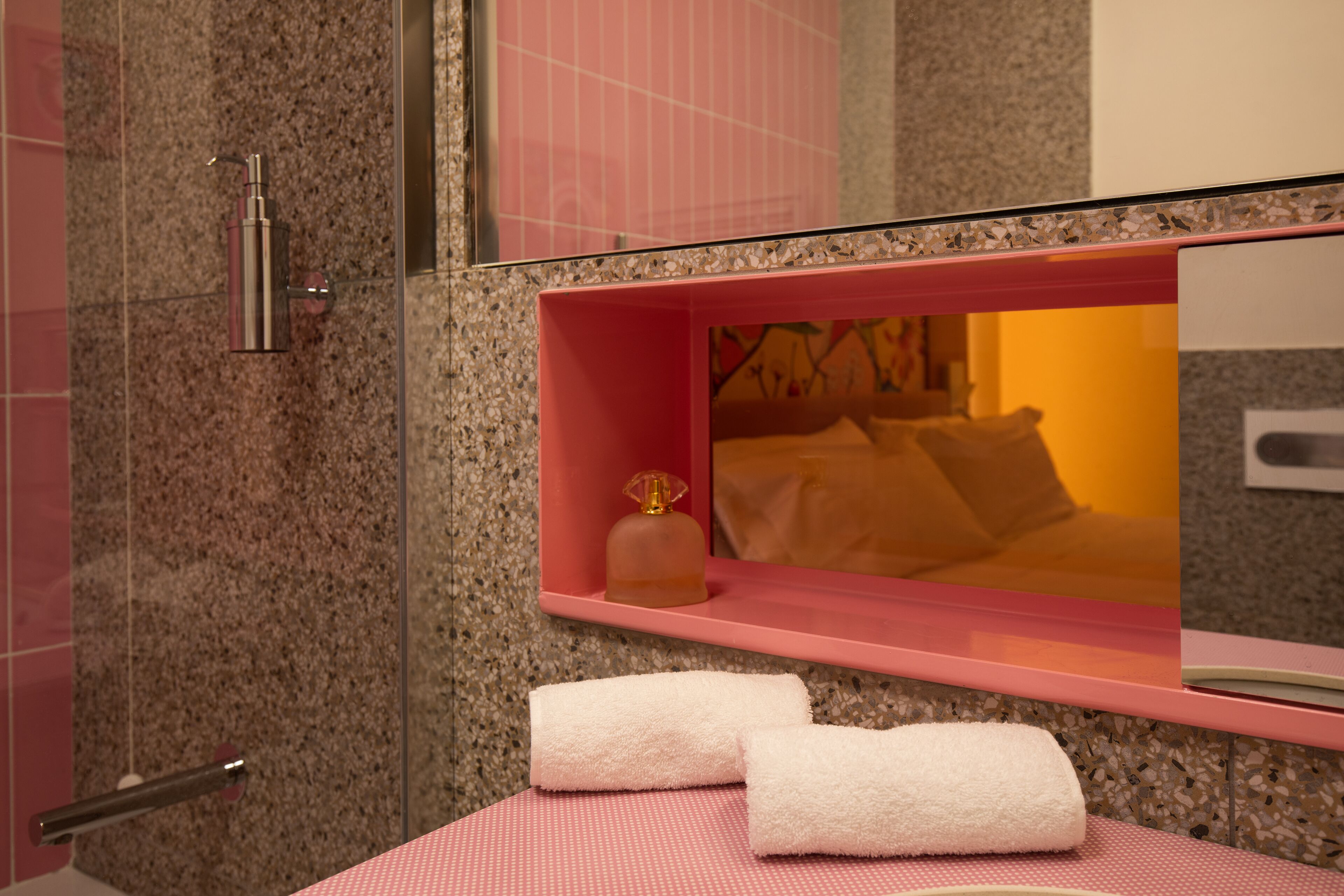 A bathroom with a granite wall, a pink shelf with toiletries, and two white towels on a pink mat.