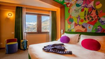 A hotel room with a bed, colorful wall art, a window with a mountain view, and a suitcase.