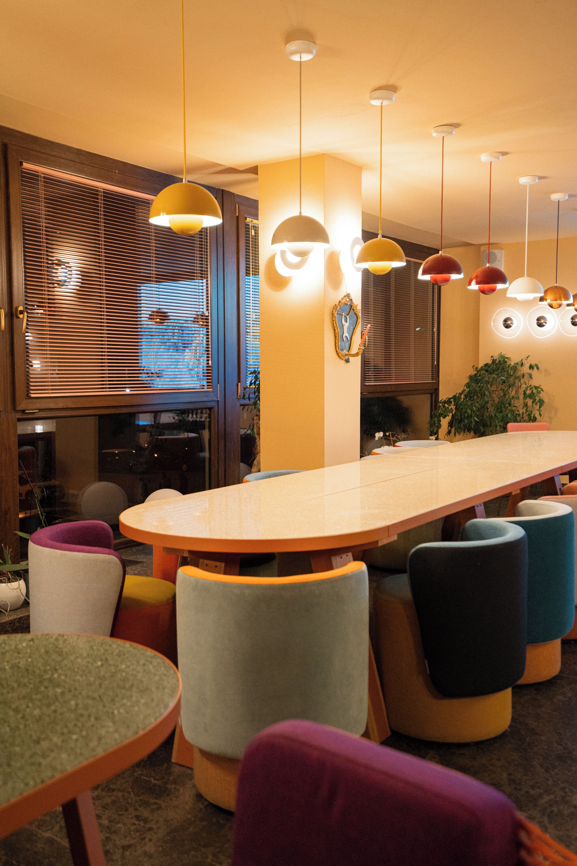 Un comedor moderno con una mesa larga y sillas coloridas, iluminación colgante y un amplio ventanal con persianas.