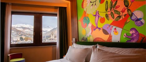 Una habitación de hotel con una cama, un mural colorido, una ventana con vistas a montañas nevadas y una silla.
