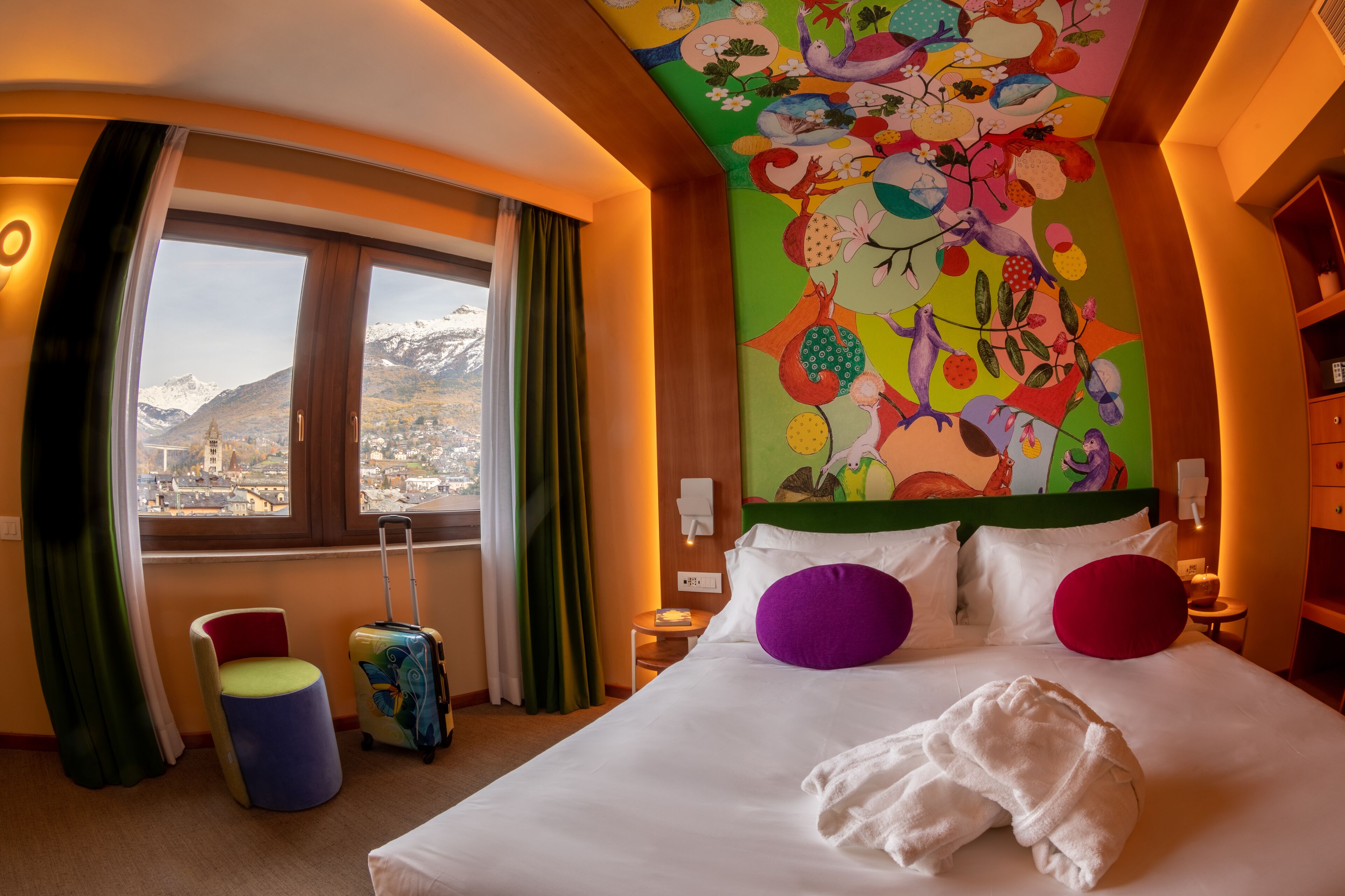 Una habitación de hotel con una cama, arte murales coloridos y vistas a montañas, además de una ventana con cortinas.
