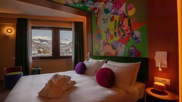 Une chambre d’hôtel avec un lit, des œuvres d’art colorées sur les murs, une vue sur les montagnes et une fenêtre avec des rideaux.