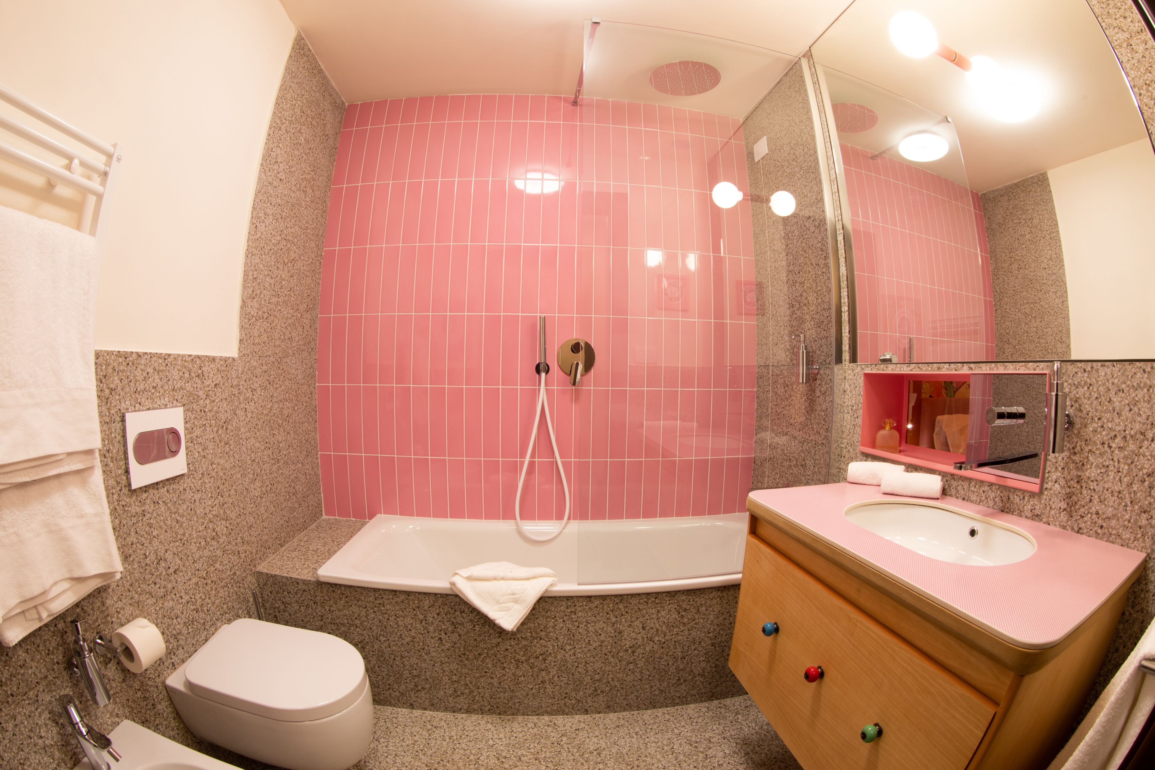 A bathroom with a pink tiled shower wall, a white bathtub, a wooden vanity with a pink sink, and a white toilet.