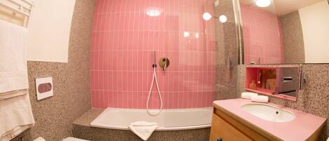 A bathroom with a pink tiled shower wall, a white bathtub, a wooden vanity with a pink sink, and a white toilet.