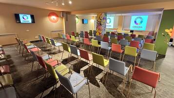 Une salle de conférence avec des rangées de chaises et de tables aux couleurs vives, un grand écran affichant le logo de l’hôtel OMAMA, et une décoration murale florale.