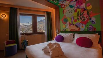 Quarto de hotel com uma cama, vista para montanhas, um mural colorido e uma janela com cortinas.