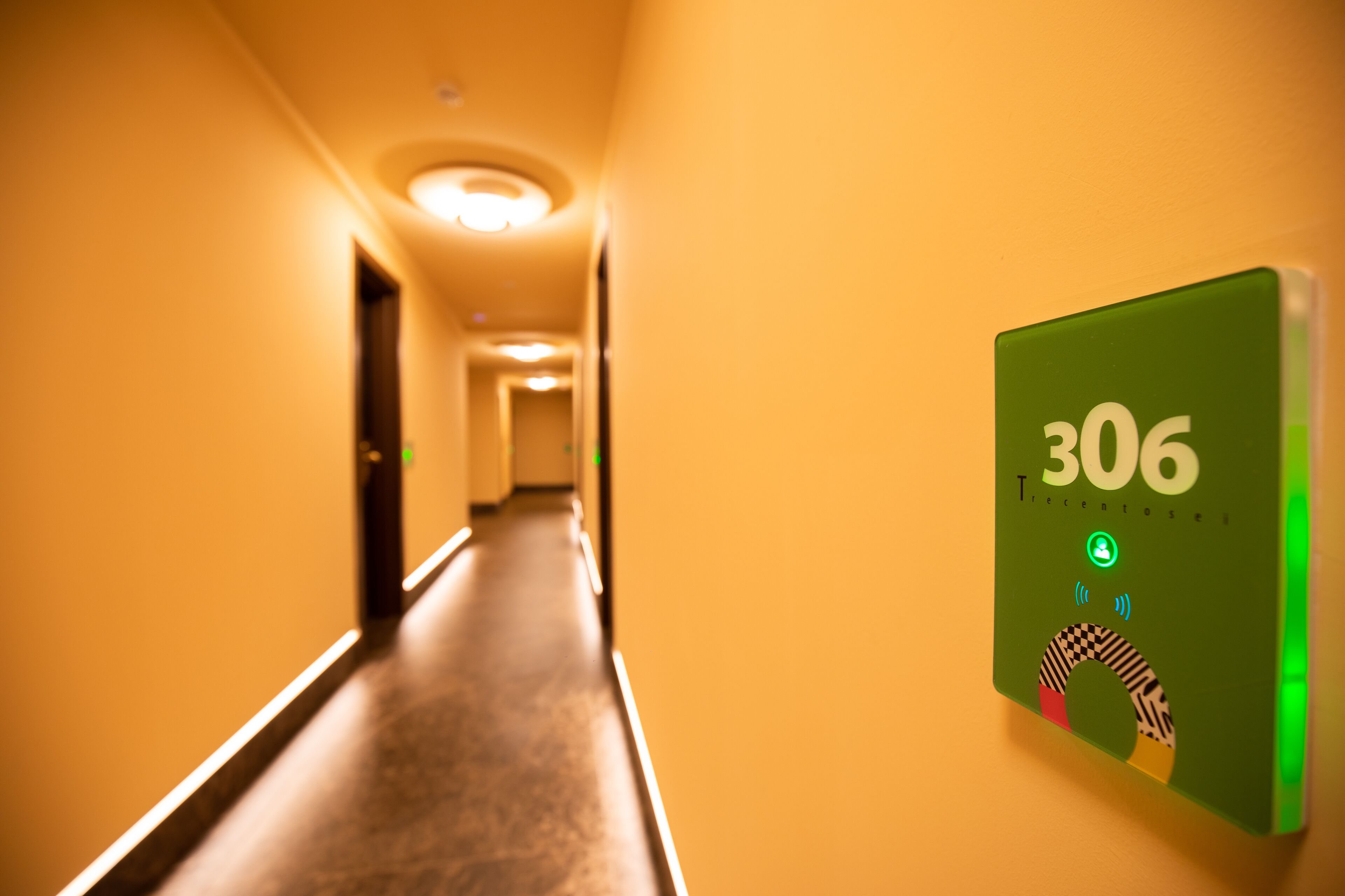 Un pasillo con un dispositivo verde fijado a la pared que muestra el número de habitación 306, un diseño de cuadros y un icono circular.