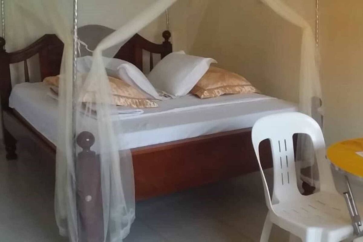 Uma cama com estrutura de madeira e dossel, uma cadeira de plástico branca e uma mesa redonda pequena com tampo amarelo.